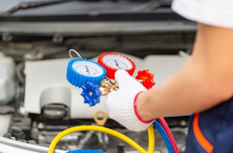A gloved mechanic holds manifold pressure gauges while servicing a car's air conditioning system.