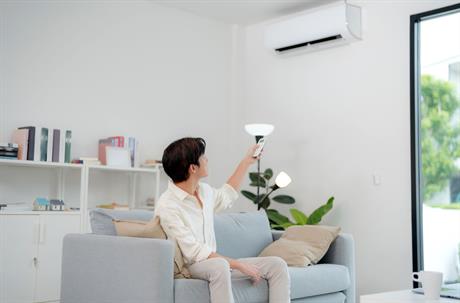 A man sitting on a sofa uses a remote control to operate a wall-mounted air conditioner.