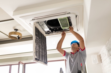 A technician in safety gear services a ceiling cassette air conditioner.
