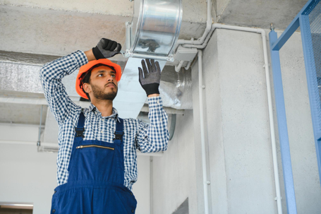 A technician wearing a hard hat installs ventilation ductwork on a ceiling.