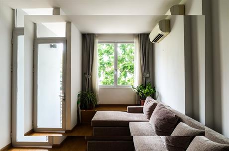 A sectional sofa sits in a bright room with a wall-mounted air conditioner and a window overlooking greenery.
