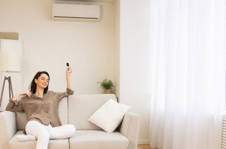 A woman relaxes on a sofa while using a remote control to operate a wall-mounted air conditioner.