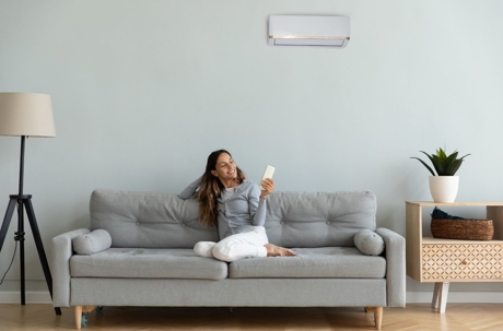 A woman relaxes on a sofa while using a remote control to operate a wall-mounted air conditioner.