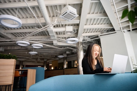 A young woman works on her laptop while sitting on a couch in a modern open-plan office space.