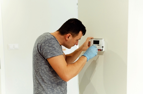 A man wearing blue gloves installs a thermostat on a wall.