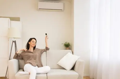 A woman relaxes on a sofa while using a remote control to operate a wall-mounted air conditioner.