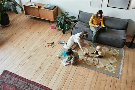 Children play with toys on a rug while an adult works on a laptop in a spacious living room.