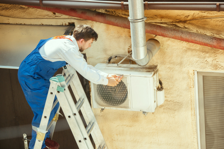 A technician standing on a ladder repairs an outdoor air conditioning unit mounted on a wall.