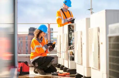 Two technicians wearing hard hats and high-visibility jackets inspect HVAC units on a rooftop.