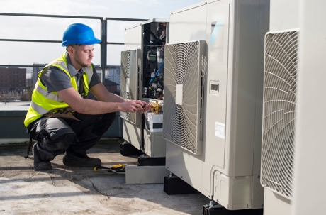 A technician wearing a hard hat and high-visibility vest services an outdoor air conditioning unit on a rooftop.