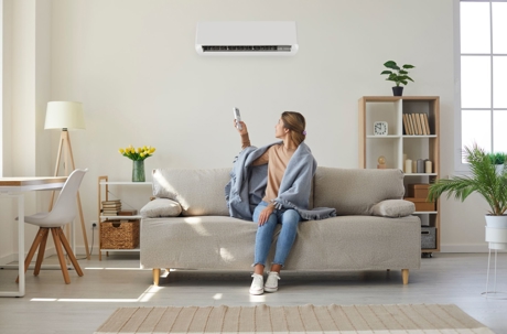 A woman relaxes on a sofa while using a remote control to operate a wall-mounted air conditioner.